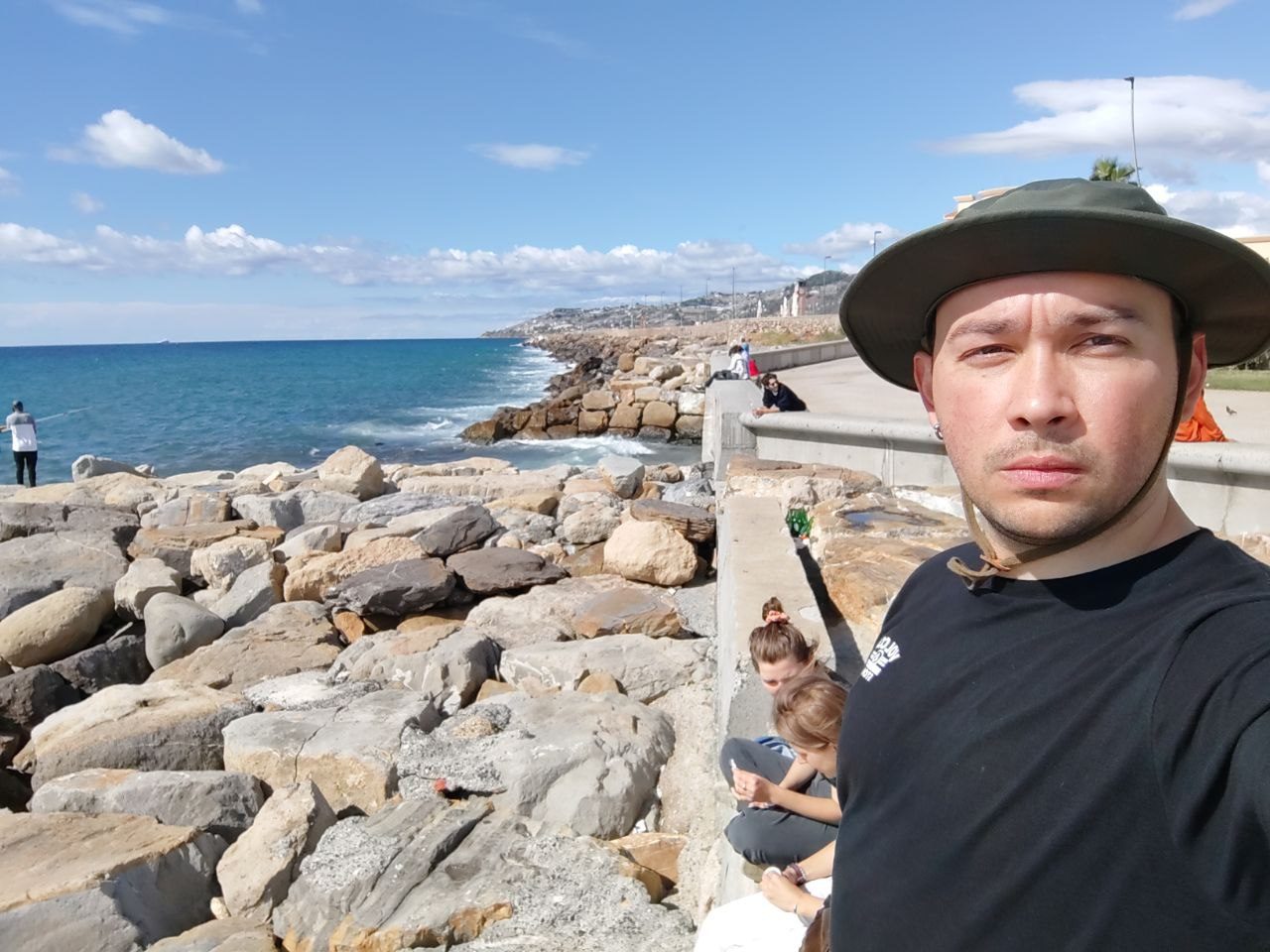 Andrew Motsyk on a seafront promenade in a wide-brimmed hat, the Mediterranean coastline behind him.